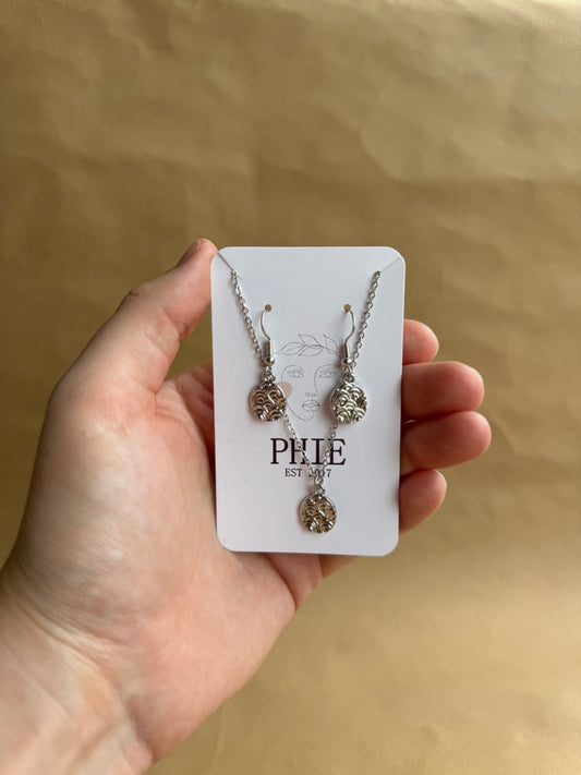 Silver necklace and earrings on a 'PHIE' card held by a hand against a beige background