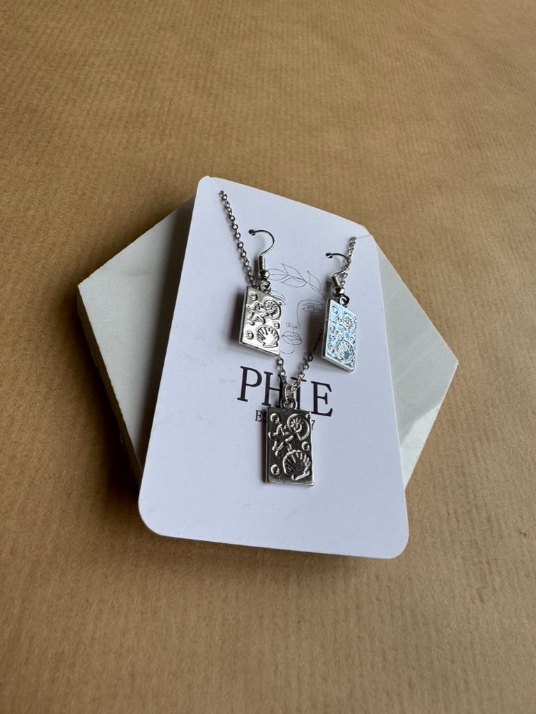 Silver jewelry set with pendant and earrings on a branded card against a beige background