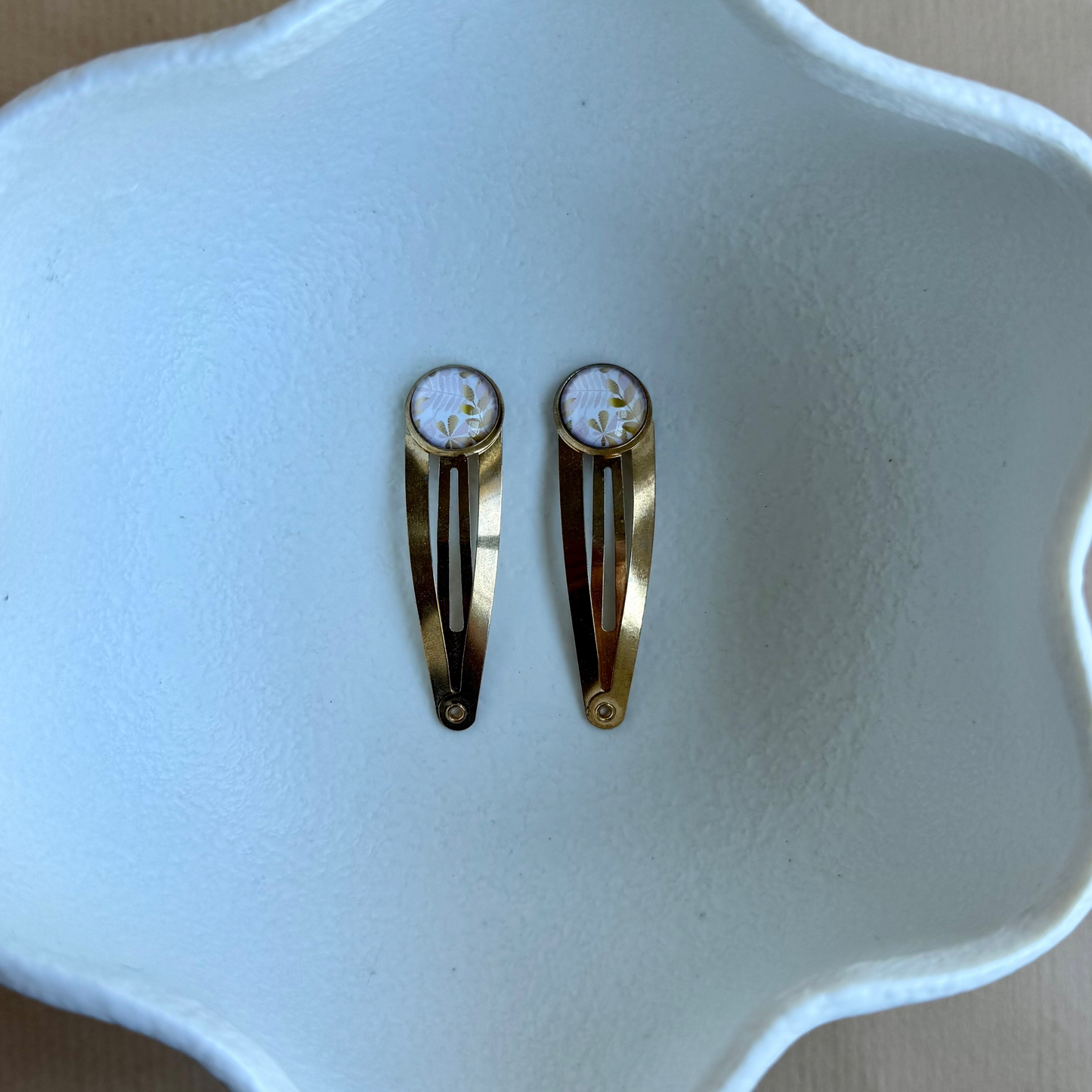 Two gold hair clips with pink and gold  stones on a white dish against a beige background