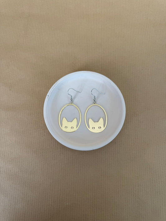 Gold cat-shaped earrings on a white circular stand against a beige background