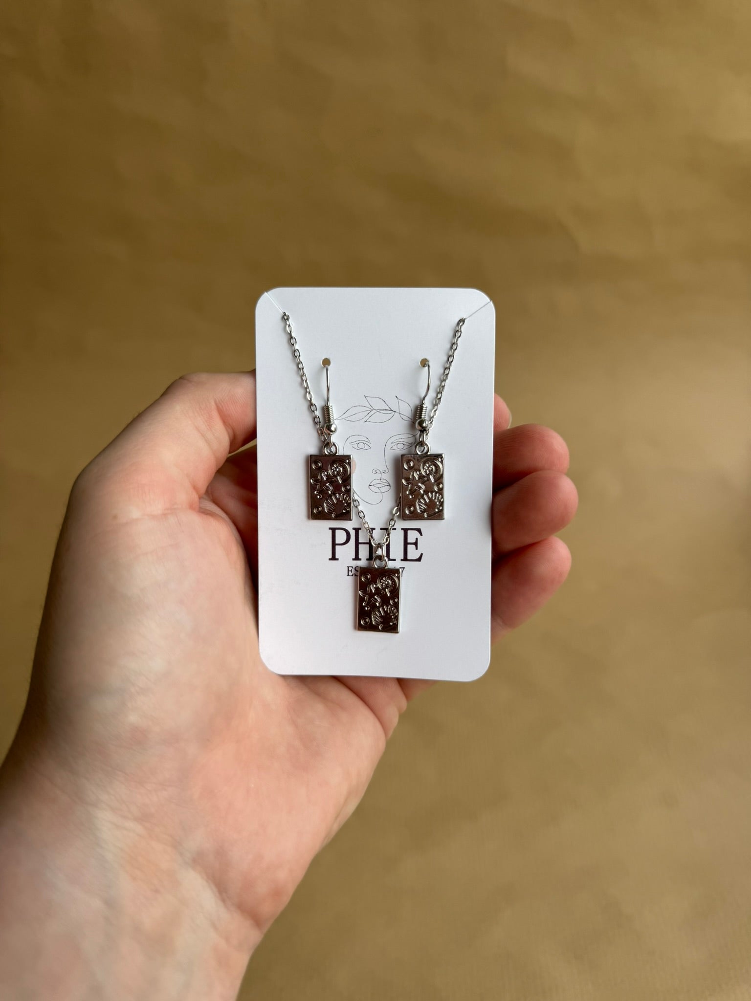 Hand holding a necklace set on a branded card against a brown background